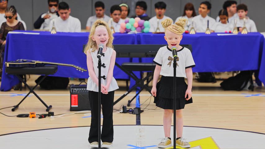 Two young girls are singing at a microphone in the middle of a gymnasium 